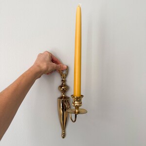 May include: A gold-colored wall sconce with a long, yellow candle. The sconce has an ornate design and a small, round plate to catch wax. The candle is tall and slender, with a lit wick.