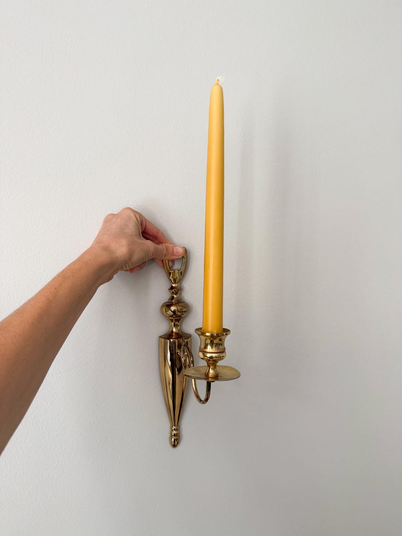 May include: A gold-colored wall sconce with a long, yellow candle. The sconce has an ornate design and a small, round plate to catch wax. The candle is tall and slender, with a lit wick.