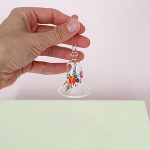 Puede incluir: Una campana de cristal transparente con un dise&ntilde;o floral en rojo, naranja y morado. La campana est&aacute; sujeta por una persona. La campana tiene un mango largo y transparente y una parte superior redondeada. El dise&ntilde;o floral est&aacute; en la parte inferior de la campana.