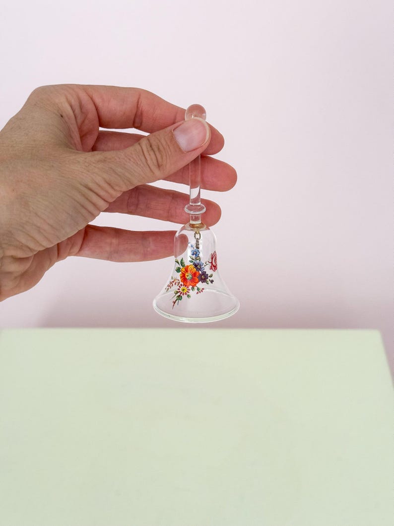 Puede incluir: Una campana de cristal transparente con un dise&ntilde;o floral en rojo, naranja y morado. La campana est&aacute; sujeta por una persona. La campana tiene un mango largo y transparente y una parte superior redondeada. El dise&ntilde;o floral est&aacute; en la parte inferior de la campana.
