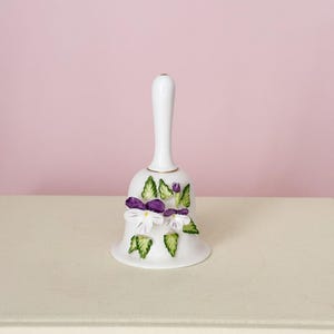 Puede incluir: Una campana de porcelana blanca con un mango largo, adornada con flores moradas y blancas y detalles de hojas verdes. La campana tiene un borde dorado. Un art&iacute;culo decorativo.