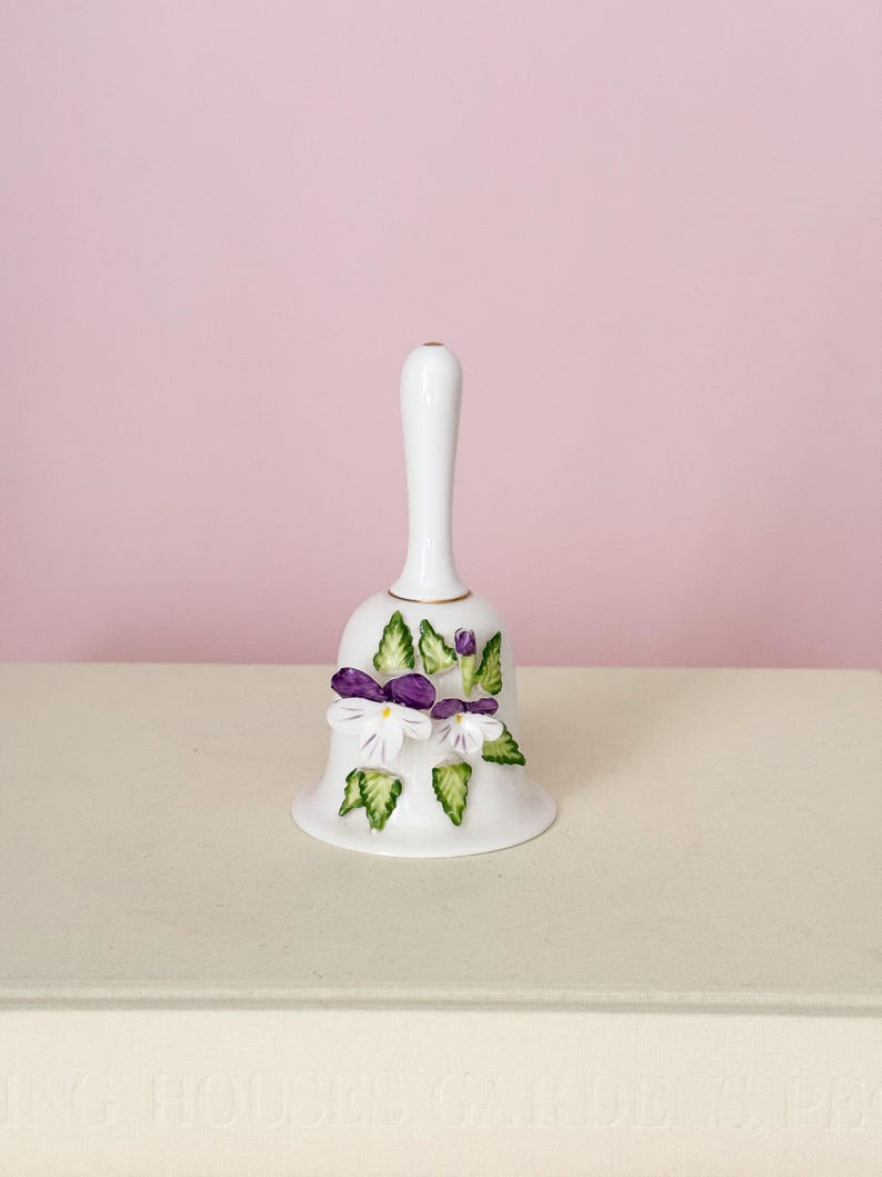 Puede incluir: Una campana de porcelana blanca con un mango largo, adornada con flores moradas y blancas y detalles de hojas verdes. La campana tiene un borde dorado. Un art&iacute;culo decorativo.