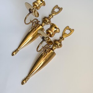 May include: A collection of three vintage brass candle holders. Each holder features a conical base, a decorative handle, and a circular plate to catch wax. The holders are a shiny gold color and are arranged on a white surface.