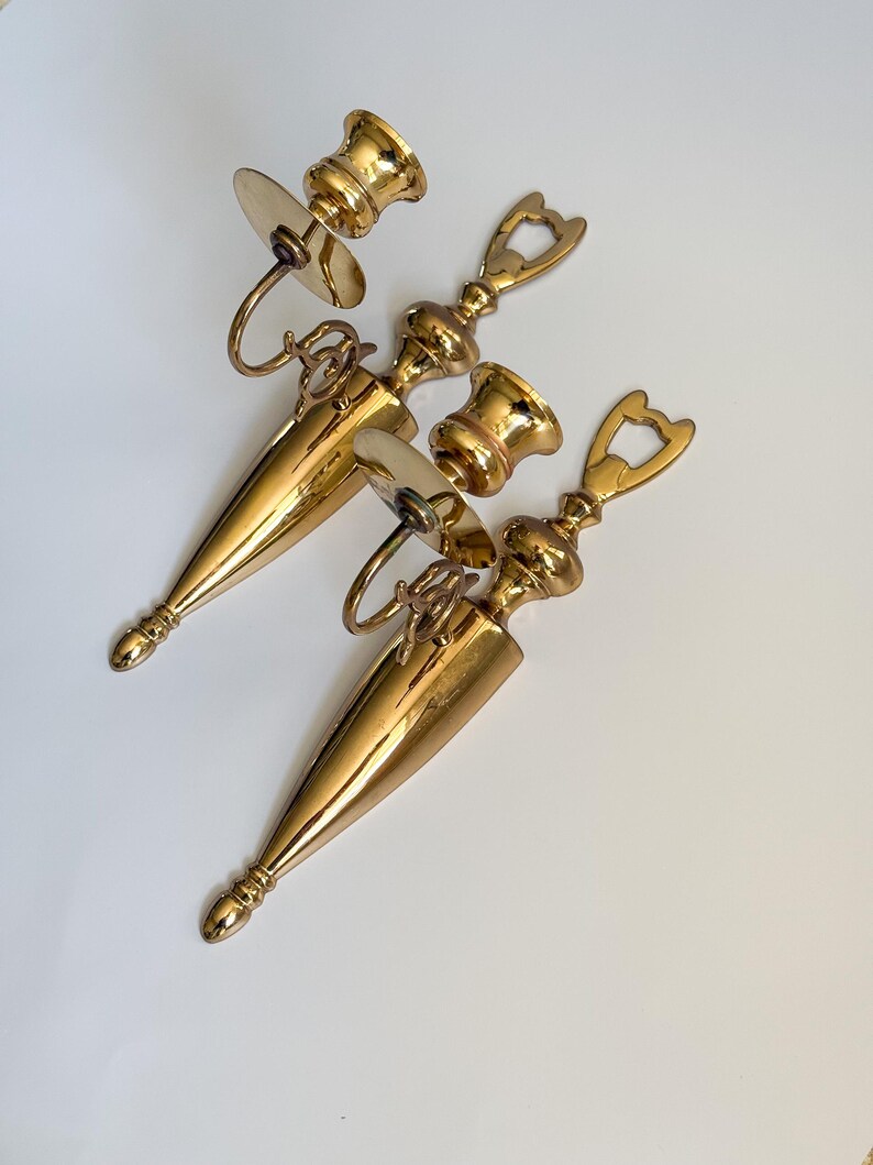 May include: A collection of three vintage brass candle holders. Each holder features a conical base, a decorative handle, and a circular plate to catch wax. The holders are a shiny gold color and are arranged on a white surface.