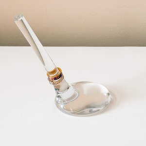 May include: A clear glass ring holder featuring a slanted, tapered post attached to a circular base. Three rings are stacked on the post: a gold band with blue and red enamel details, a thin gold band, and a gold band with small clear stones.