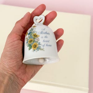 Puede incluir: Campana de cer&aacute;mica blanca con asa en forma de coraz&oacute;n y dise&ntilde;o floral. La campana presenta el texto "A Mother is the heart of home" en escritura azul. Flores amarillas y azules decoran la superficie de la campana, creando una pieza decorativa encantadora.