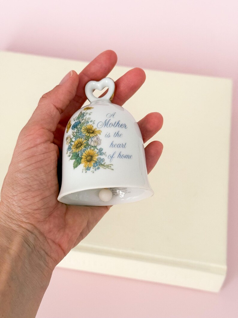 Puede incluir: Campana de cer&aacute;mica blanca con asa en forma de coraz&oacute;n y dise&ntilde;o floral. La campana presenta el texto "A Mother is the heart of home" en escritura azul. Flores amarillas y azules decoran la superficie de la campana, creando una pieza decorativa encantadora.