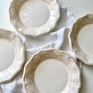 May include: Four white ceramic plates with a scalloped edge and a beige floral pattern border. The plates are arranged on a white linen cloth, creating a classic and elegant aesthetic. The plates are ideal for serving meals.