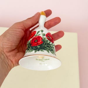 Puede incluir: Campana de cer&aacute;mica blanca con asa dorada y dise&ntilde;o floral de amapolas. La campana presenta amapolas rojas con hojas verdes y el texto "The Summer Collection London". Hecho en Inglaterra.
