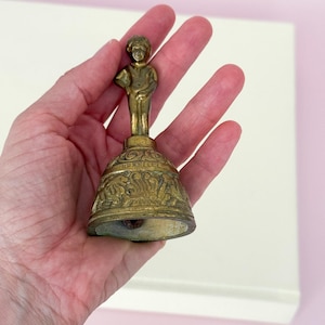 Puede incluir: Una campana de lat&oacute;n vintage con una peque&ntilde;a figura encima de una base decorativa. La campana tiene un aspecto envejecido y antiguo, con intrincados detalles alrededor de la base. La figura es una peque&ntilde;a estatua de un ni&ntilde;o. La campana se sostiene en una mano.