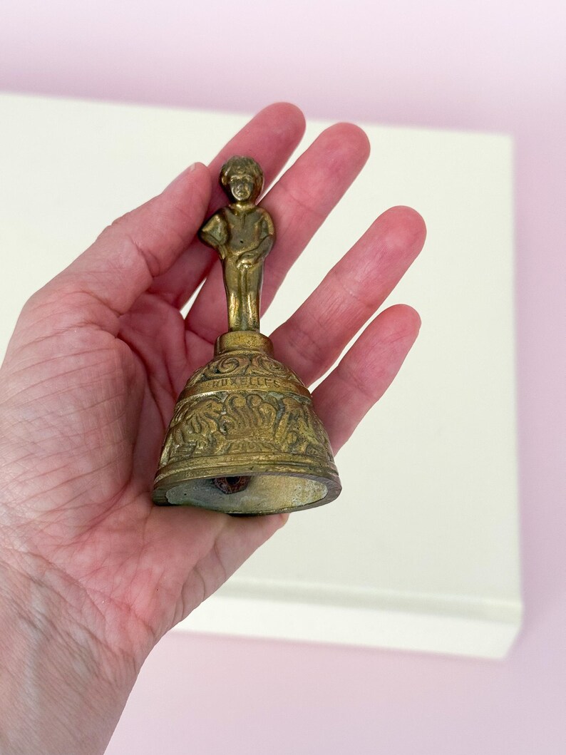 Puede incluir: Una campana de lat&oacute;n vintage con una peque&ntilde;a figura encima de una base decorativa. La campana tiene un aspecto envejecido y antiguo, con intrincados detalles alrededor de la base. La figura es una peque&ntilde;a estatua de un ni&ntilde;o. La campana se sostiene en una mano.