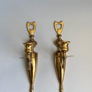 May include: Two ornate, polished brass wall sconces. Each sconce features a candle holder, a small shelf, and a decorative top. The sconces have a teardrop shape and are mounted on a white wall. The brass has a shiny, reflective finish.