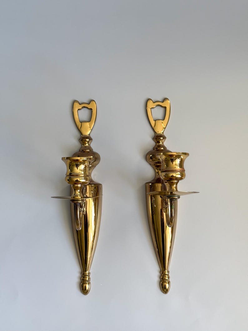 May include: Two ornate, polished brass wall sconces. Each sconce features a candle holder, a small shelf, and a decorative top. The sconces have a teardrop shape and are mounted on a white wall. The brass has a shiny, reflective finish.