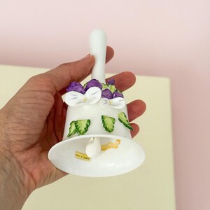 Puede incluir: Campana de cer&aacute;mica blanca con asa, adornada con dise&ntilde;os de flores moradas y blancas y hojas verdes. La campana tiene una base acampanada y un peque&ntilde;o badajo en el interior. La campana se sostiene en una mano.