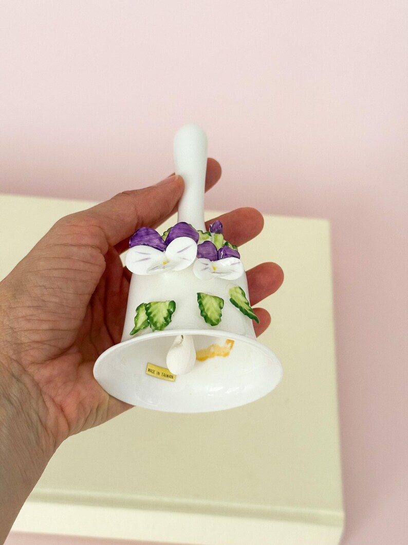 Puede incluir: Campana de cer&aacute;mica blanca con asa, adornada con dise&ntilde;os de flores moradas y blancas y hojas verdes. La campana tiene una base acampanada y un peque&ntilde;o badajo en el interior. La campana se sostiene en una mano.