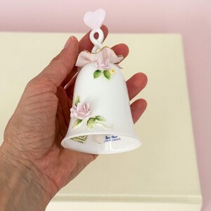 Puede incluir: Campana de porcelana blanca con detalles de rosas rosas, un lazo rosa y un asa en forma de coraz&oacute;n. La campana tiene ribetes dorados y hojas verdes. El texto "Best China 1979 ENESCO" es visible.
