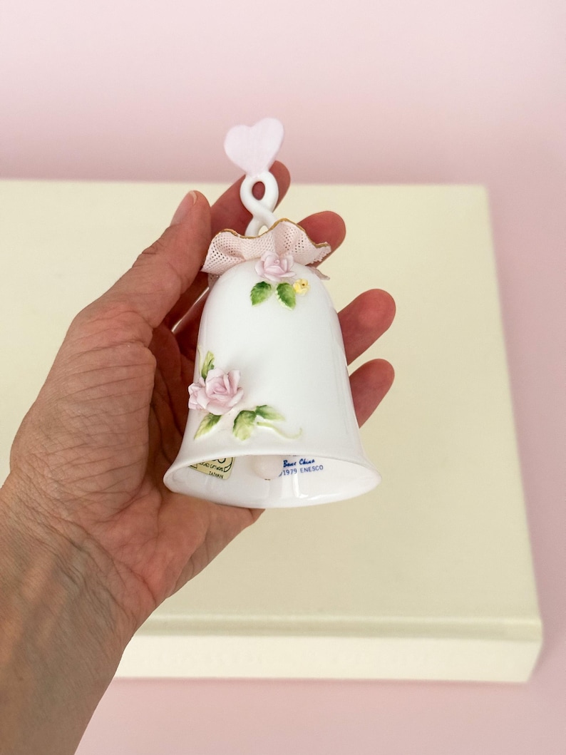 Puede incluir: Campana de porcelana blanca con detalles de rosas rosas, un lazo rosa y un asa en forma de coraz&oacute;n. La campana tiene ribetes dorados y hojas verdes. El texto "Best China 1979 ENESCO" es visible.