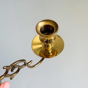 May include: A vintage brass candlestick holder with a decorative arm. The candlestick has a round base and a cup-shaped holder for the candle. The brass has a polished, shiny finish.