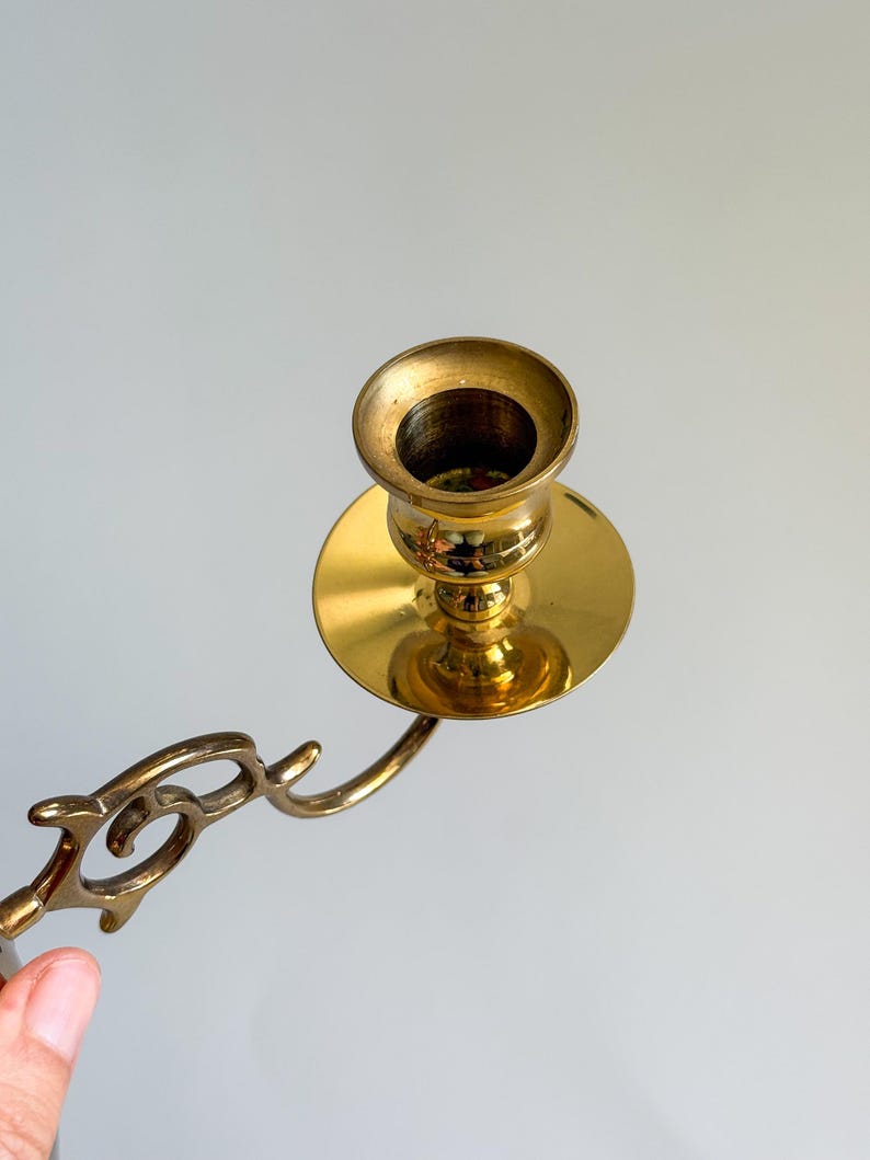 May include: A vintage brass candlestick holder with a decorative arm. The candlestick has a round base and a cup-shaped holder for the candle. The brass has a polished, shiny finish.