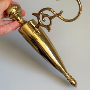 May include: A gold-colored, vintage-style wall sconce with a decorative, ornate design. The sconce features a conical body, a curved arm, and an elaborate, swirling accent. The sconce is made of metal and has a polished finish.