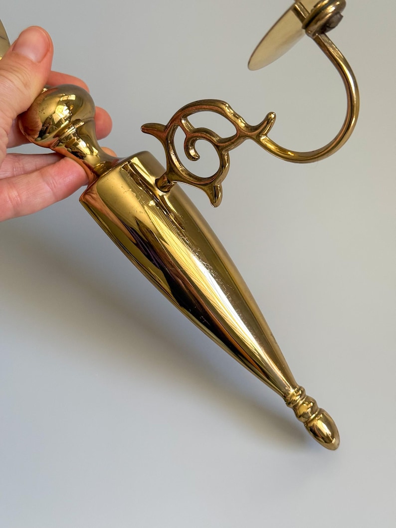 May include: A gold-colored, vintage-style wall sconce with a decorative, ornate design. The sconce features a conical body, a curved arm, and an elaborate, swirling accent. The sconce is made of metal and has a polished finish.