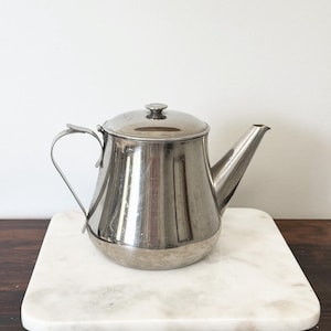 May include: A polished stainless steel teapot with a rounded base, a curved handle, a tapered spout, and a matching lid with a small knob. The metal surface is reflective and shows subtle signs of use, featuring a classic, functional design.