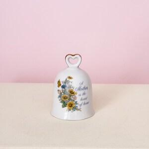 Puede incluir: Campana de cer&aacute;mica blanca con asa en forma de coraz&oacute;n y dise&ntilde;o floral con flores amarillas y azules y una mariposa. La campana tiene el texto "A Mother is the heart of home".