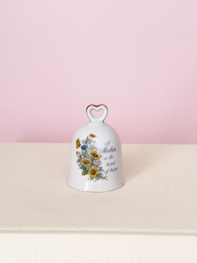Puede incluir: Campana de cer&aacute;mica blanca con asa en forma de coraz&oacute;n y dise&ntilde;o floral con flores amarillas y azules y una mariposa. La campana tiene el texto "A Mother is the heart of home".