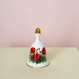 Puede incluir: Campana de cer&aacute;mica blanca con asa y borde dorados. La campana presenta un dise&ntilde;o floral de amapolas rojas con hojas verdes. La campana est&aacute; sobre una superficie blanca sobre un fondo rosa.