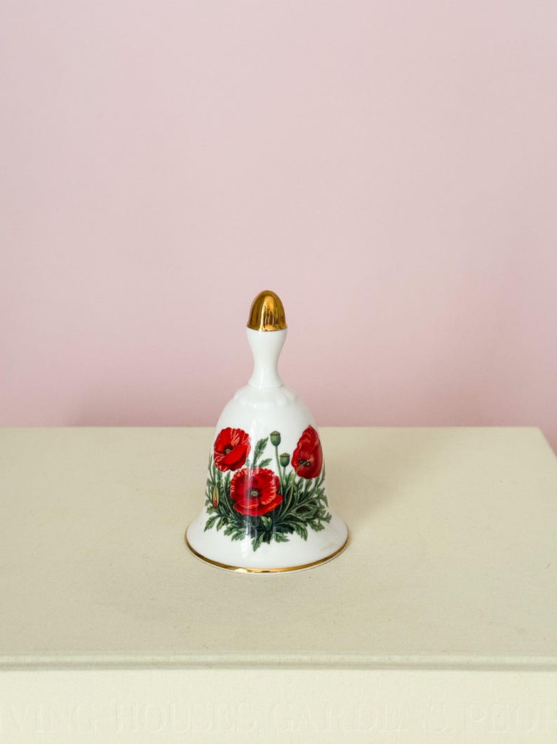 Puede incluir: Campana de cer&aacute;mica blanca con asa y borde dorados. La campana presenta un dise&ntilde;o floral de amapolas rojas con hojas verdes. La campana est&aacute; sobre una superficie blanca sobre un fondo rosa.