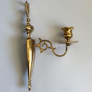 May include: A vintage brass wall sconce with a decorative design. The sconce features a candle holder and an ornate arm with a scroll design. The sconce is made of polished brass and is designed to be mounted on a wall. The sconce is approximately 12 inches in height.