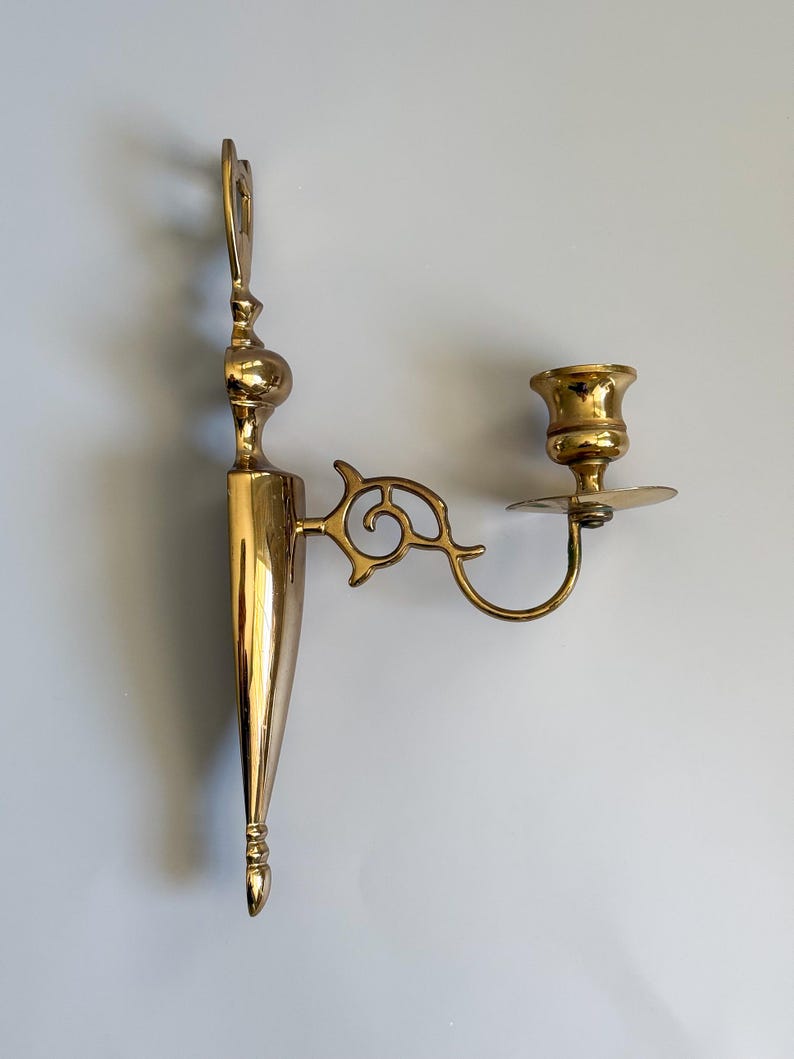 May include: A vintage brass wall sconce with a decorative design. The sconce features a candle holder and an ornate arm with a scroll design. The sconce is made of polished brass and is designed to be mounted on a wall. The sconce is approximately 12 inches in height.