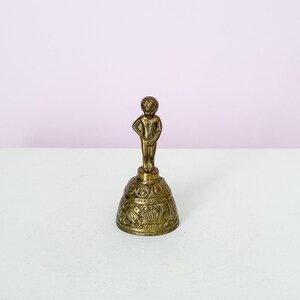 Puede incluir: Una peque&ntilde;a campana de lat&oacute;n antiguo con una figura de un ni&ntilde;o. La campana tiene detalles ornamentados alrededor de la base y la figura del ni&ntilde;o est&aacute; de pie con una mano en la cadera. Un objeto decorativo.