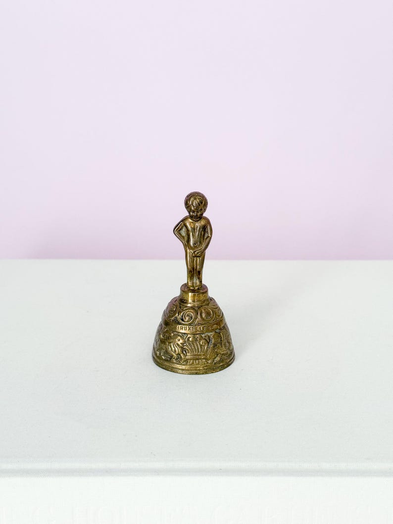 Puede incluir: Una peque&ntilde;a campana de lat&oacute;n antiguo con una figura de un ni&ntilde;o. La campana tiene detalles ornamentados alrededor de la base y la figura del ni&ntilde;o est&aacute; de pie con una mano en la cadera. Un objeto decorativo.