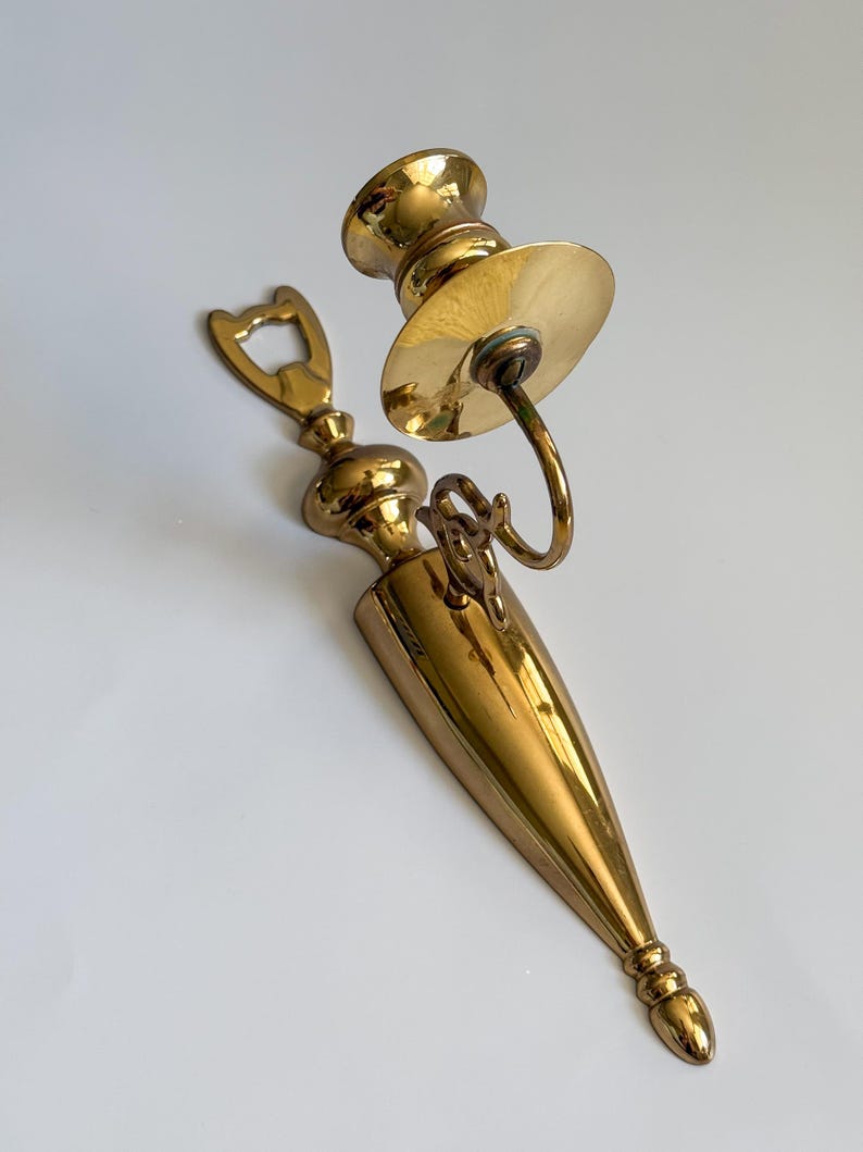 May include: A vintage brass candle holder and bottle opener set. The candle holder features a circular base and a curved arm. The bottle opener has a decorative top. Both items have a polished finish.