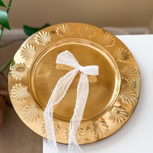 May include: A gold-colored decorative plate with a scalloped edge design. A white, sheer ribbon with small white polka dots is tied in a bow and rests on the plate. The plate is on a white surface.