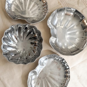 May include: Four silver-colored, seashell-shaped dishes. Each dish has a unique design, with textured surfaces and scalloped edges. The dishes are arranged on a neutral-toned fabric, creating a coastal or nautical theme.