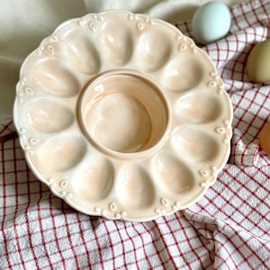 Op de afbeelding: Een crèmekleurige keramische eierserveerschaal met een centrale kom en 12 eierinkepingen. De schaal heeft een geschulpte rand met decoratieve details. De achtergrond omvat een rood-wit geruite doek en verschillende eieren.