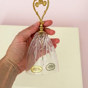 Puede incluir: Una campana de cristal transparente con un asa dorada en forma de coraz&oacute;n. La campana tiene un dise&ntilde;o facetado y una peque&ntilde;a bola de cristal en el interior. El asa est&aacute; adornada con detalles florales. La campana tiene dos pegatinas que dicen "GARANTIZADO ORO DE 24 QUILATES" y "Genuine LERO CRYSTAL".