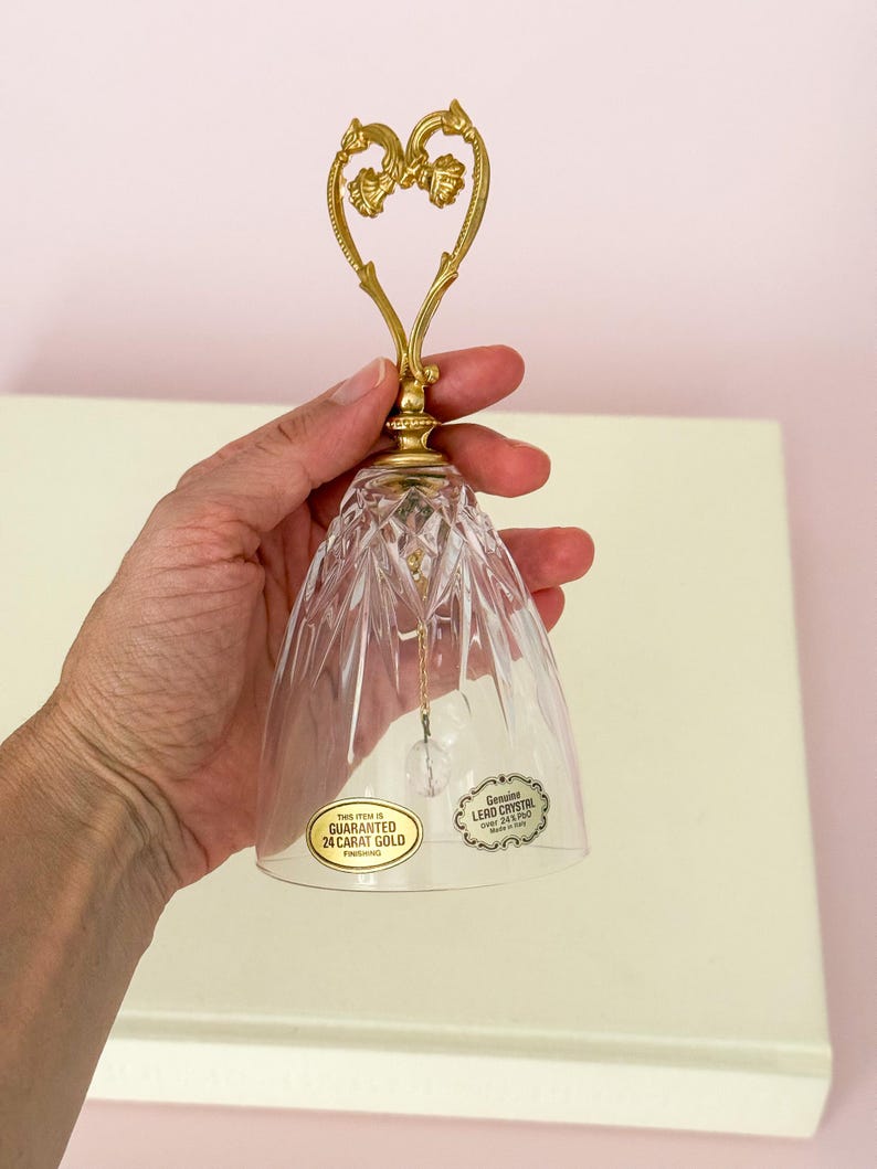 Puede incluir: Una campana de cristal transparente con un asa dorada en forma de coraz&oacute;n. La campana tiene un dise&ntilde;o facetado y una peque&ntilde;a bola de cristal en el interior. El asa est&aacute; adornada con detalles florales. La campana tiene dos pegatinas que dicen "GARANTIZADO ORO DE 24 QUILATES" y "Genuine LERO CRYSTAL".
