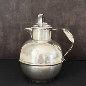 Puede incluir: Una tetera o cafetera esférica plateada con asa y tapa. La jarra tiene un pico para verter y un cuerpo redondeado. El asa es curva y está unida al lateral. La tapa tiene un asa pequeña.
