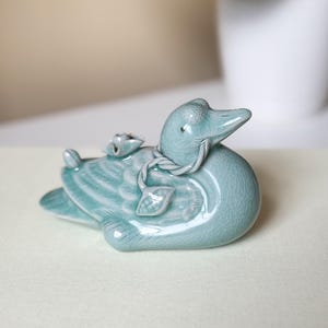 Puede incluir: Figura de pato de cerámica azul claro con acabado craquelado. El pato tiene alas detalladas y un elemento decorativo en la espalda. Es un artículo decorativo.