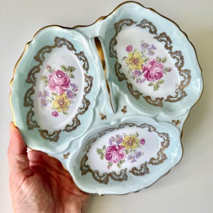 May include: A three-sectioned, light blue porcelain serving dish with a gold trim. Each section features a floral design with pink and yellow roses. The dish has a handle and is likely used for serving snacks or appetizers.