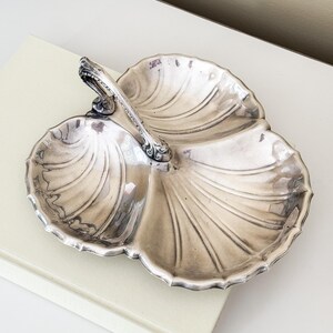 May include: Silver-toned, three-sectioned serving dish with a handle. The dish has a scalloped edge and a seashell-like design. The handle is ornate with decorative detailing. The dish is resting on a light-colored surface.