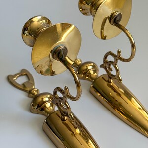 May include: Two vintage brass wall sconces. Each sconce features a circular plate, a curved arm, and a conical base. The brass has a polished finish, reflecting light. The sconces are designed to hold candles, adding a touch of elegance to any space.
