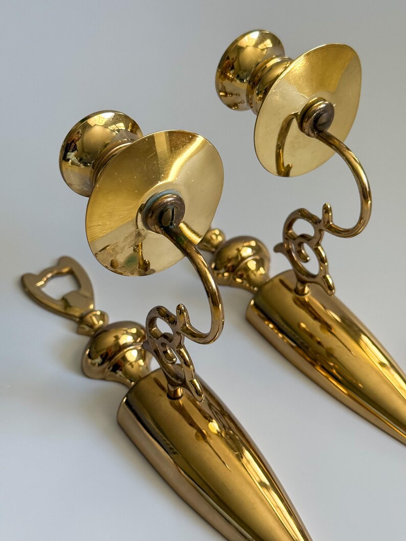 May include: Two vintage brass wall sconces. Each sconce features a circular plate, a curved arm, and a conical base. The brass has a polished finish, reflecting light. The sconces are designed to hold candles, adding a touch of elegance to any space.