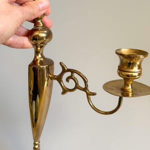 May include: A vintage brass wall sconce with a candle holder. The sconce has an ornate design with a long, tapered body and a curved arm holding the candle cup. The finish is a shiny gold color.