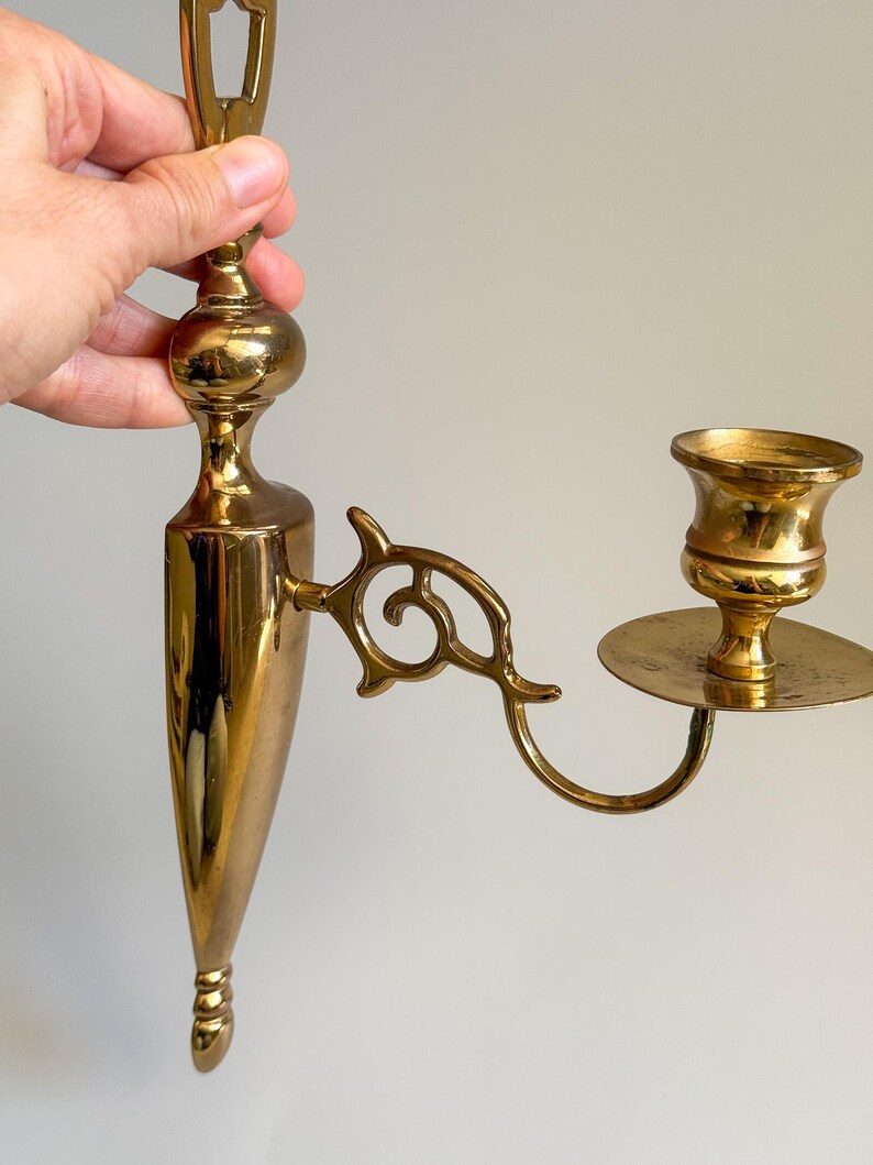 May include: A vintage brass wall sconce with a candle holder. The sconce has an ornate design with a long, tapered body and a curved arm holding the candle cup. The finish is a shiny gold color.