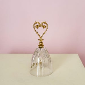 Puede incluir: Una campana de cristal transparente con un asa dorada en forma de coraz&oacute;n. La campana tiene un dise&ntilde;o tallado y se apoya sobre una superficie blanca sobre un fondo rosa. El asa presenta detalles florales ornamentados.