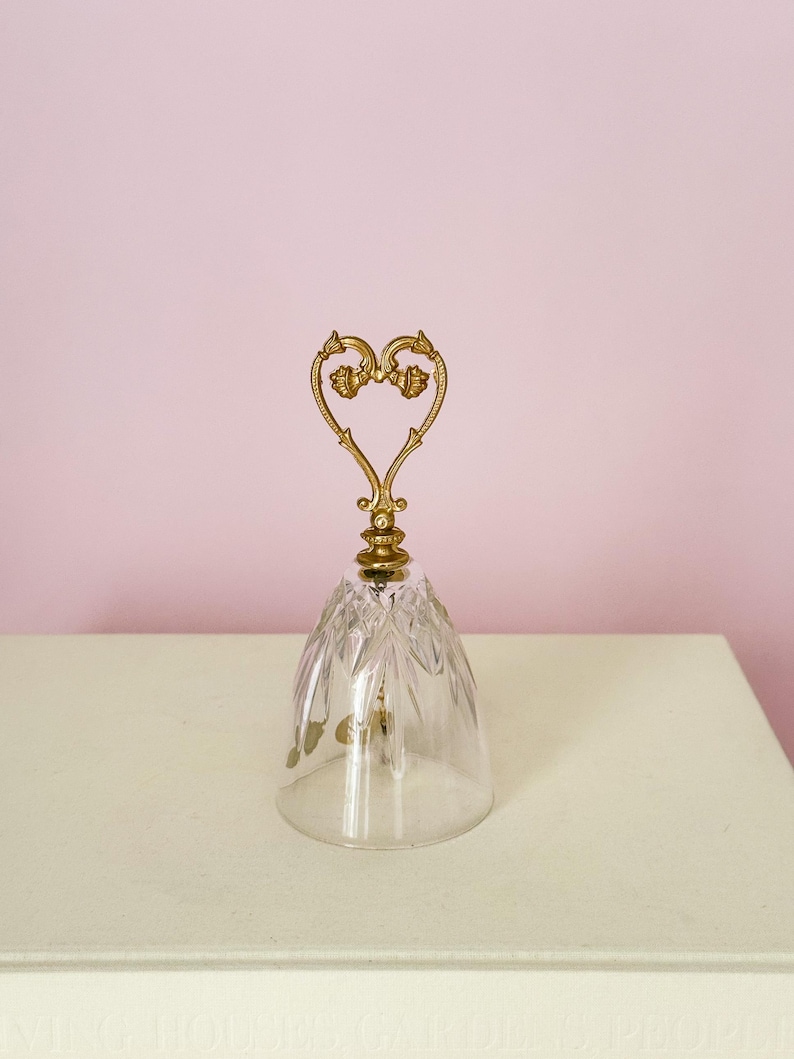 Puede incluir: Una campana de cristal transparente con un asa dorada en forma de coraz&oacute;n. La campana tiene un dise&ntilde;o tallado y se apoya sobre una superficie blanca sobre un fondo rosa. El asa presenta detalles florales ornamentados.